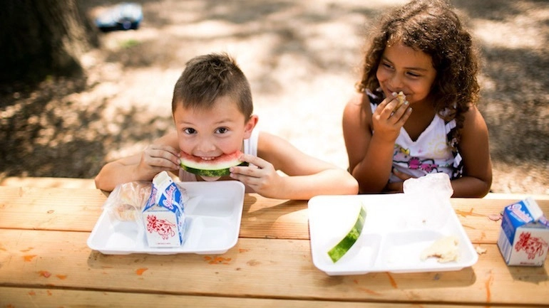 Two kids sit at a table, one eats watermelon and one laughs